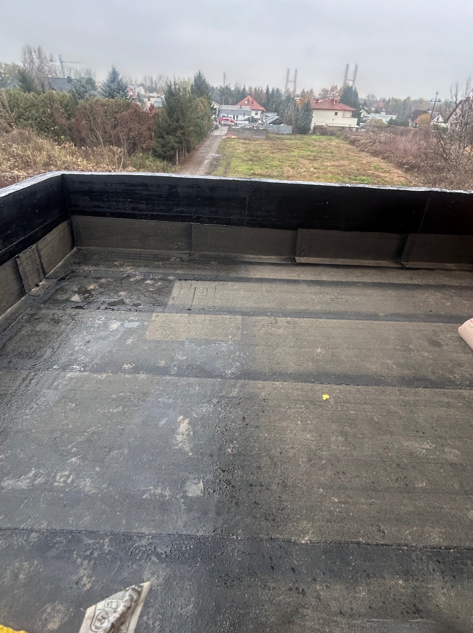 Widok z góry na płaski dach pokryty papą, z widocznymi łączeniami i elementami wykończeniowymi. W tle zabudowa jednorodzinna i drzewa pod pochmurnym niebem.