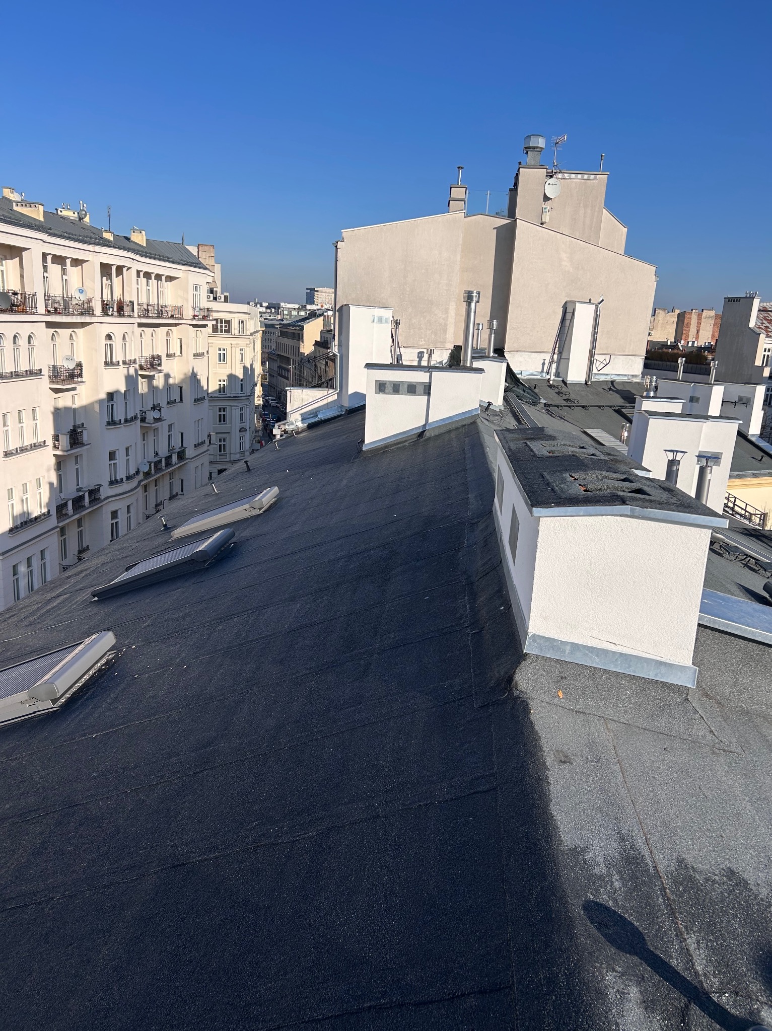 Czarny, płaski dach pokryty papą termozgrzewalną z lukarnami i kominami na tle miejskiej zabudowy w słoneczny dzień. Widok z góry.