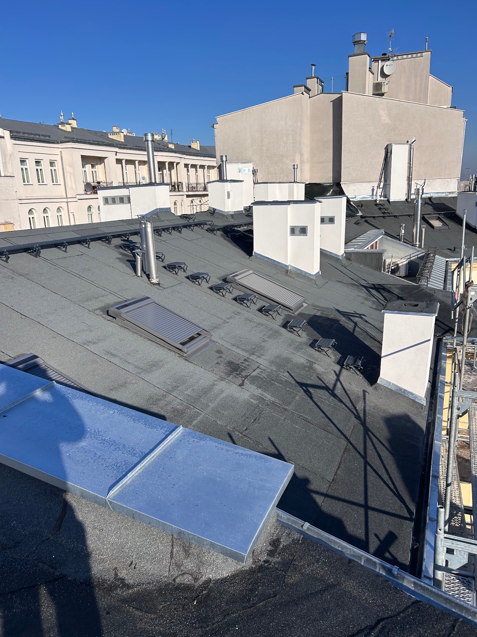 Czarny dach pokryty papą termozgrzewalną z lukarnami i kominami wentylacyjnymi na tle jasnych budynków w słoneczny dzień. Widoczne elementy rusztowania.