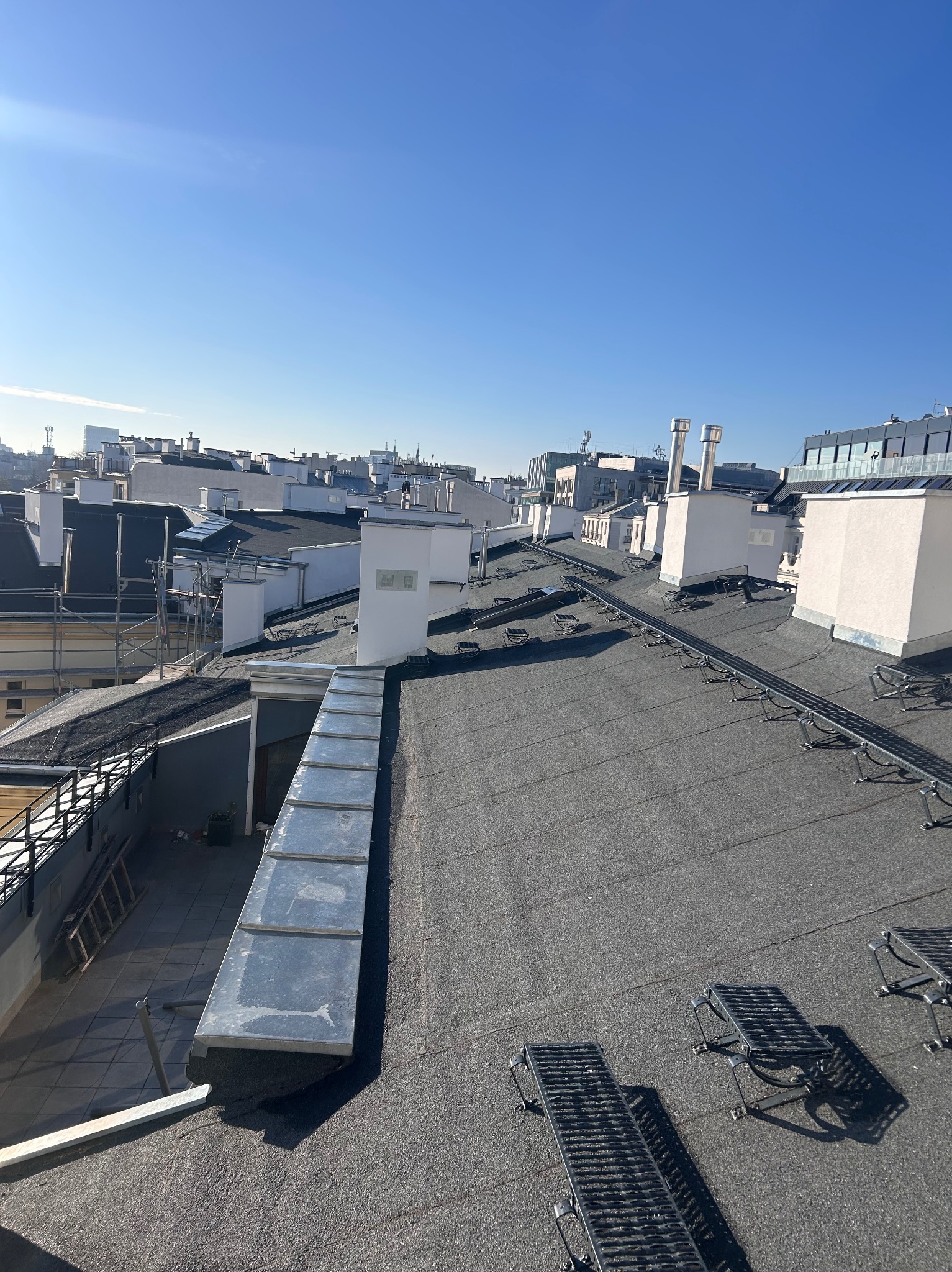 Czarny, płaski dach budynku z widocznymi elementami wentylacyjnymi i systemem komunikacji dachowej. W tle zabudowa miejska pod błękitnym niebem.