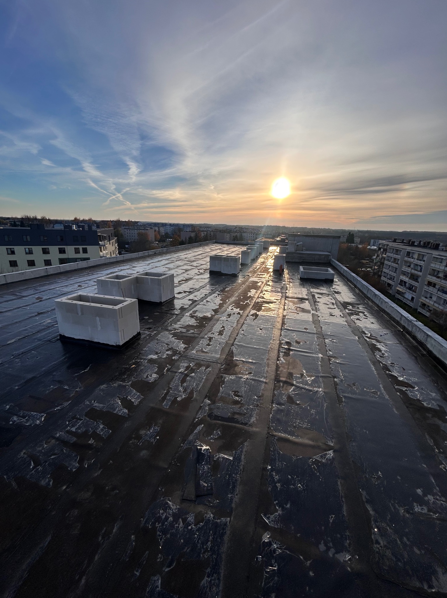 Widok z góry na płaski dach pokryty czarną papą, z betonowymi elementami, w tle zabudowa miejska i zachodzące słońce. Widoczne ślady po łatach na dachu.