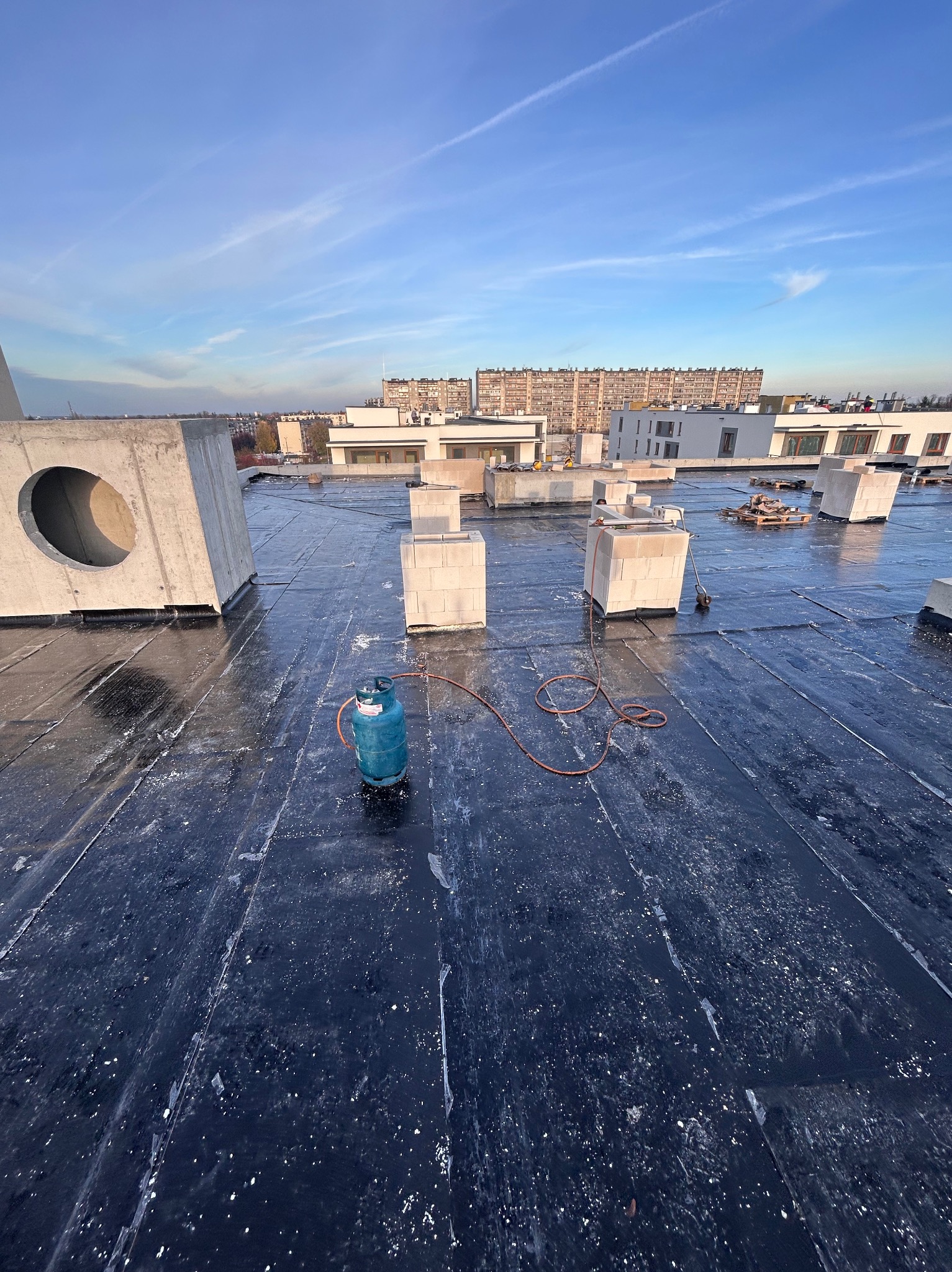 Czarna, płaska powierzchnia dachu z widocznymi elementami konstrukcyjnymi, butlą gazową i budynkami w tle, w słoneczny dzień. Widok z góry.