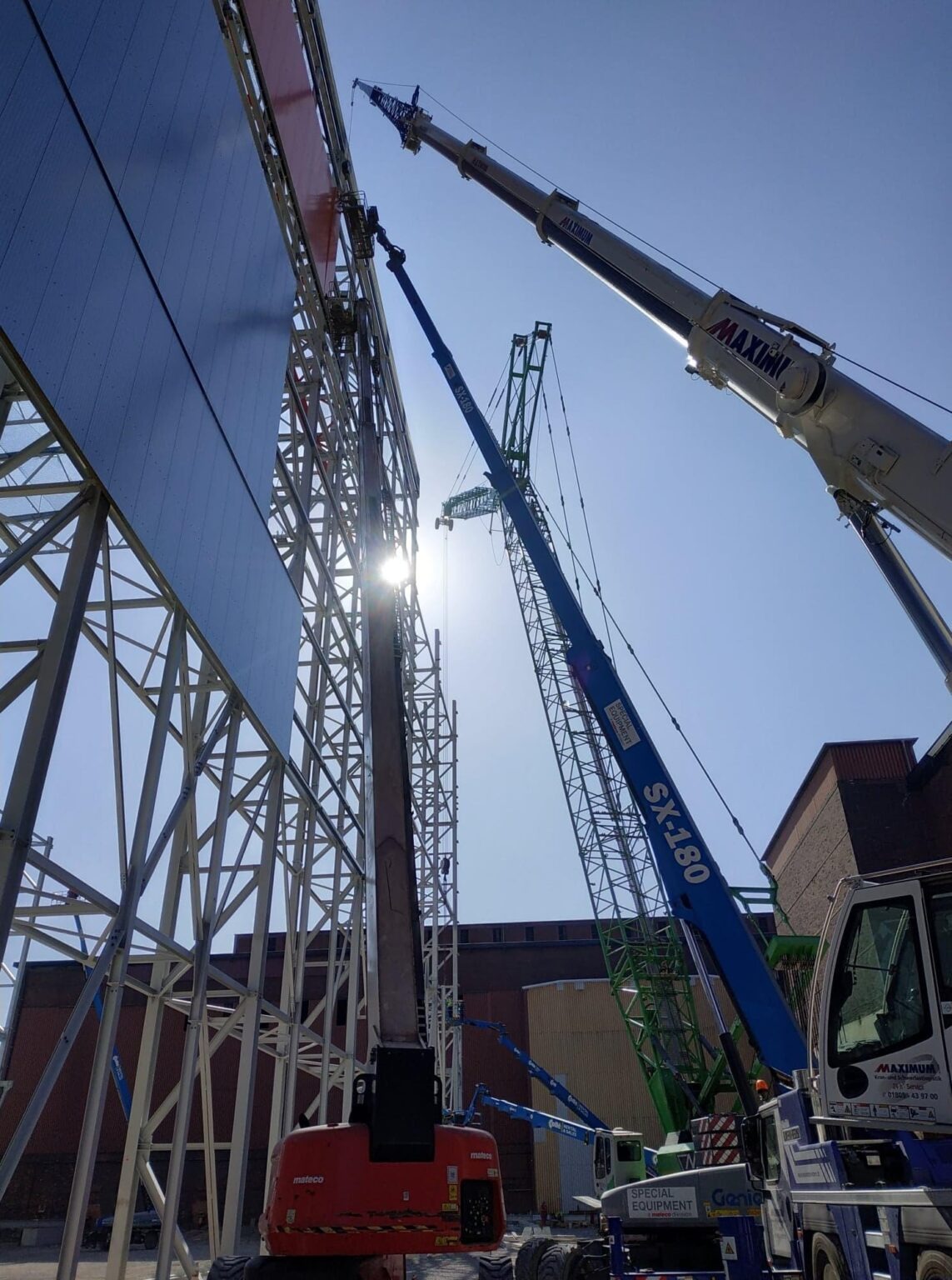 Montaż elewacji hali stalowej z użyciem dźwigów i podnośników koszowych, widok z dołu na konstrukcję i sprzęt budowlany w promieniach słońca.