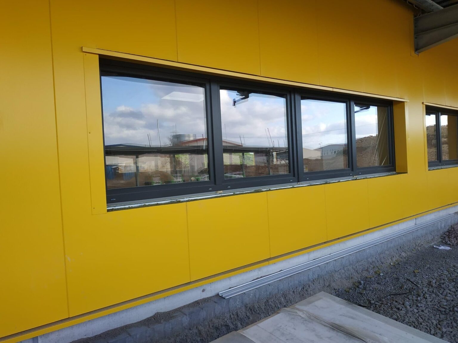 Żółta ściana hali z zamontowanymi oknami, widoczne płyty warstwowe i fundament z drobnym gruzem.