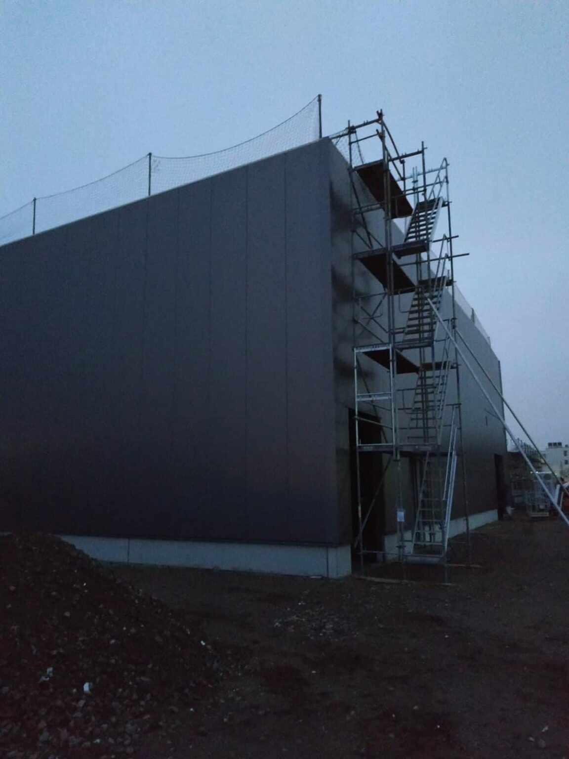 Budowa hali w trakcie: widok zewnętrzny, ściany z ciemnych płyt, rusztowania z metalowymi schodami, zabezpieczenie siatką na dachu, ziemia wokół.