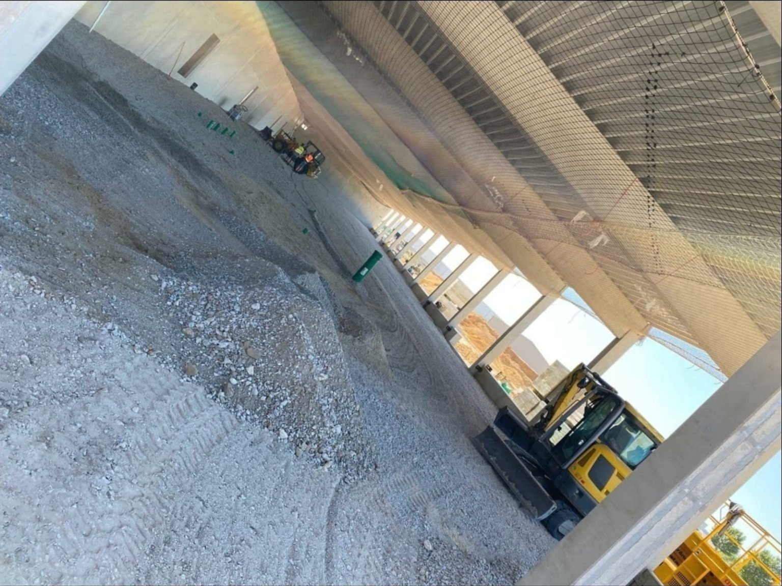 Wnętrze w trakcie budowy hali z widoczną konstrukcją stalową, siatką zabezpieczającą pod dachem i małą koparką przygotowującą podłoże, widoczne ślady opon na gruzie.