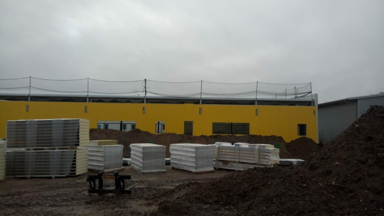 Żółta hala w trakcie budowy z przygotowanymi do montażu płytami warstwowymi na paletach, widoczna siatka zabezpieczająca na dachu i sterta ziemi.