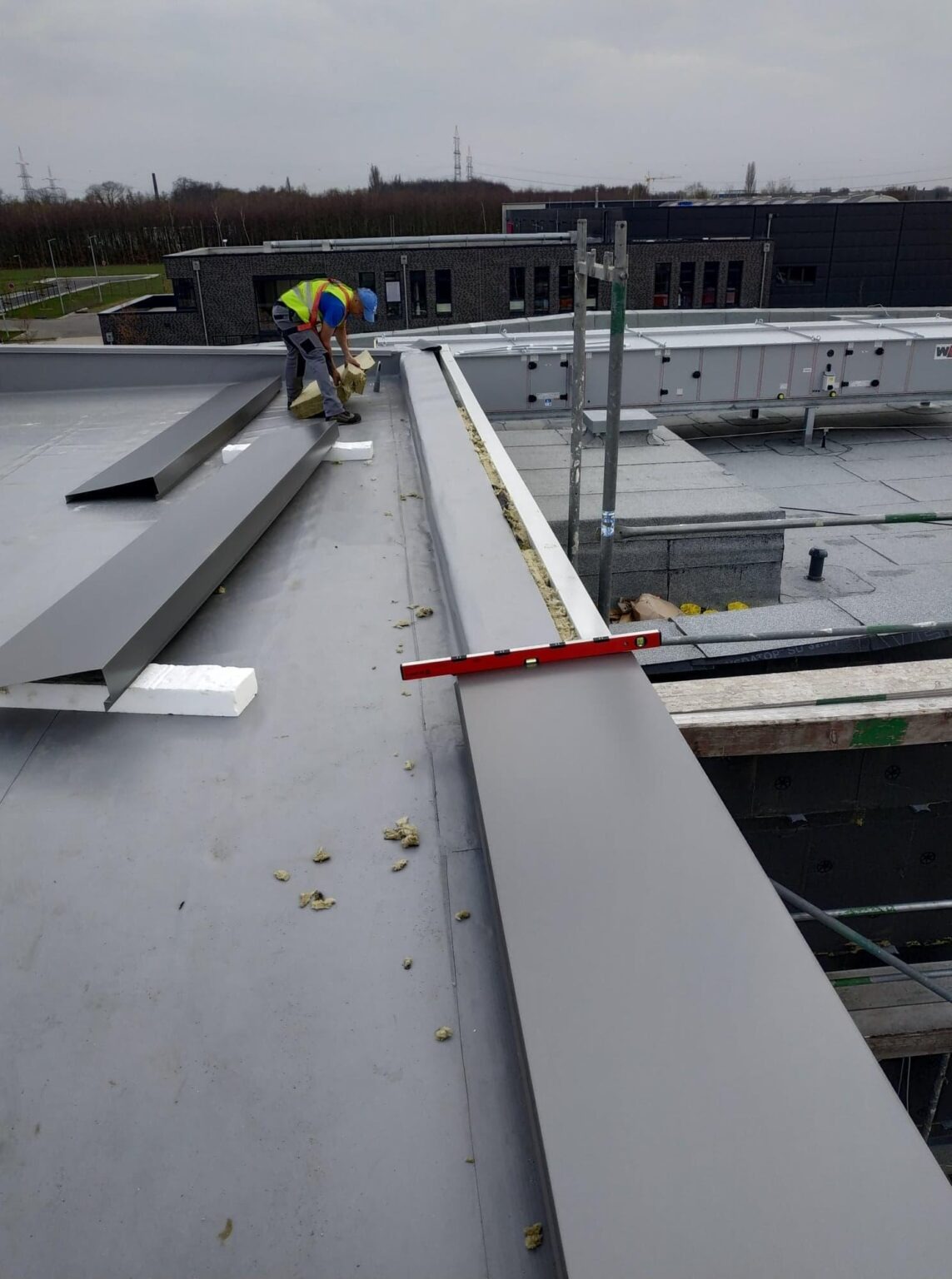 Pracownik w kamizelce odblaskowej układa izolację termiczną w metalowej obudowie na dachu budynku przemysłowego, poziomica leży na krawędzi obudowy.