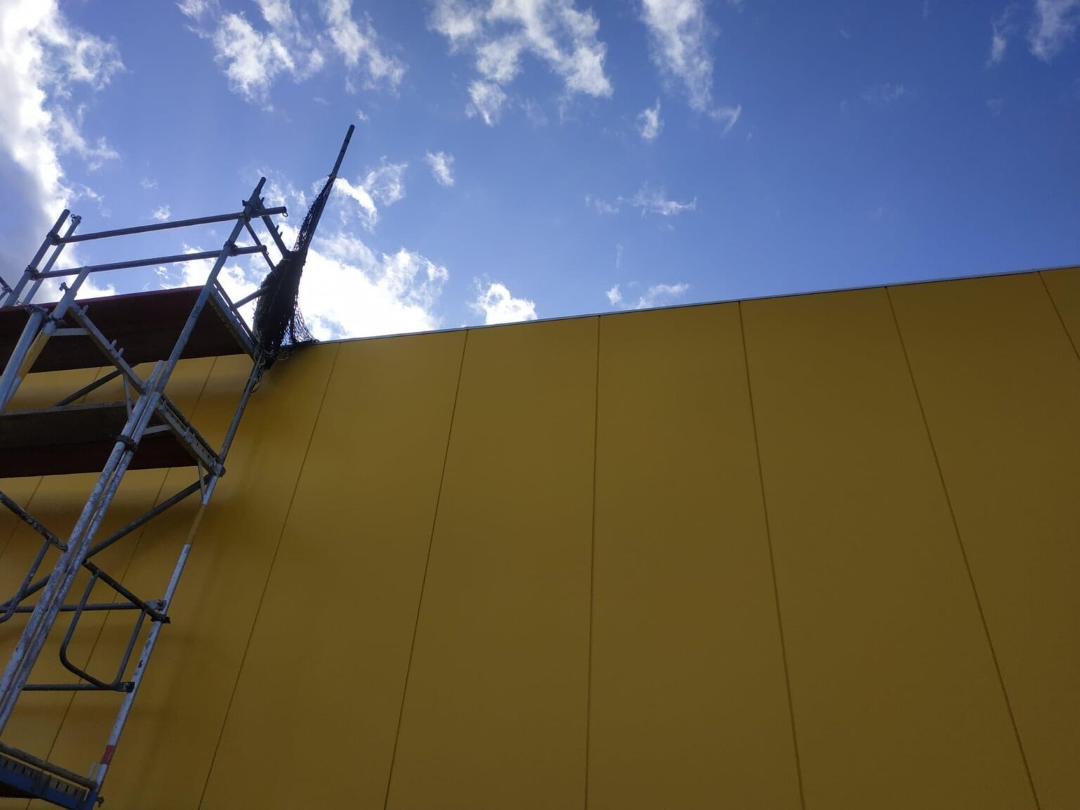 Żółta ściana hali przemysłowej, widoczna z dołu z fragmentem rusztowania i błękitnym niebem z chmurami w tle.