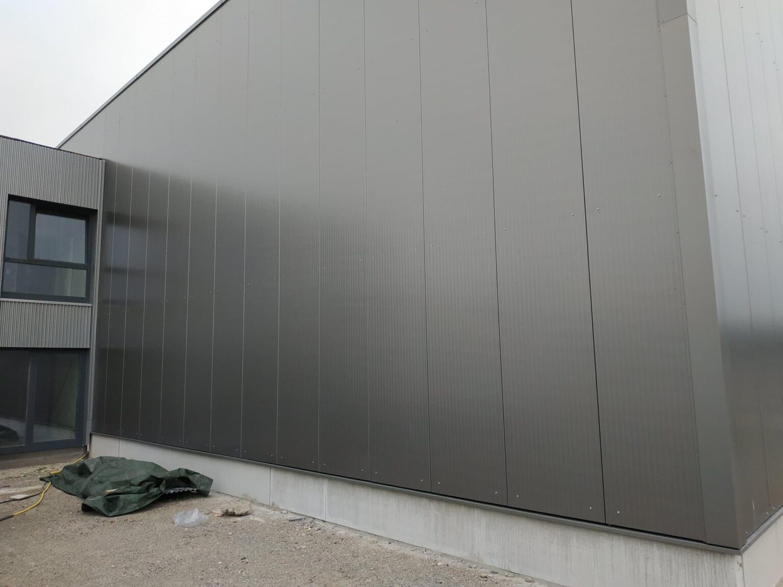 Szara ściana hali przemysłowej z płyt warstwowych, widoczne łączenia paneli i fragment okna, na ziemi plandeka i kabel.