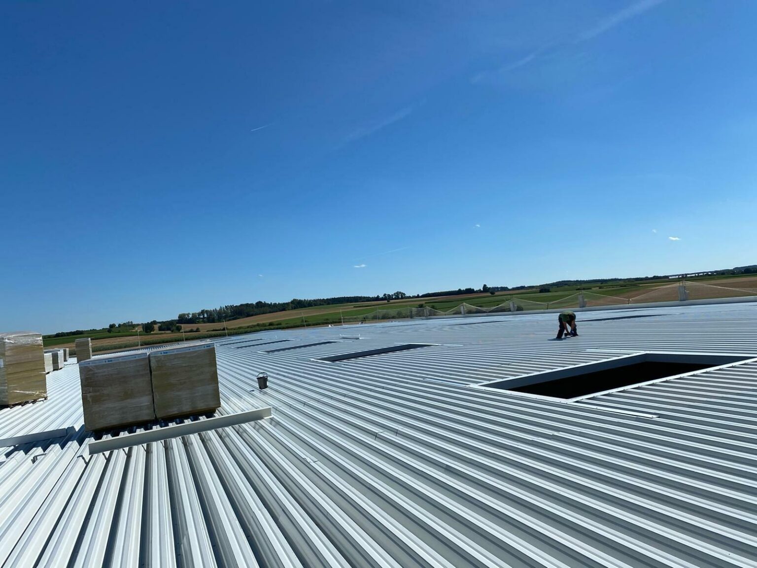 Widok z góry na rozległy dach hali z blachy trapezowej, gdzie jeden pracownik pracuje przy otworze, a w tle widać pola i lasy pod błękitnym niebem.