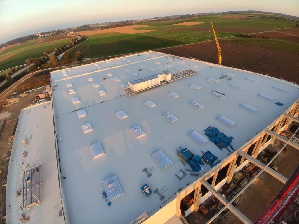 Widok z lotu ptaka na budowę hali z białym dachem z świetlikami, w tle pola uprawne i las, widoczny dźwig budowlany.