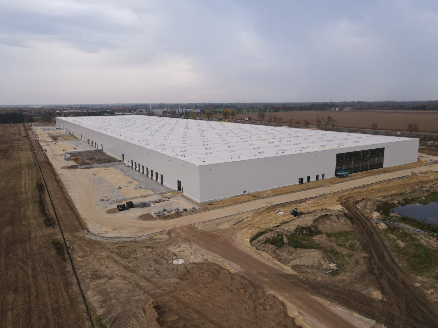 Szeroki kadr z drona na budowę dużej, białej hali magazynowej w Platerowie, z widocznymi przygotowanymi fundamentami, częściowo zamontowaną szklaną fasadą i rozległym, jesiennym krajobrazem w tle.