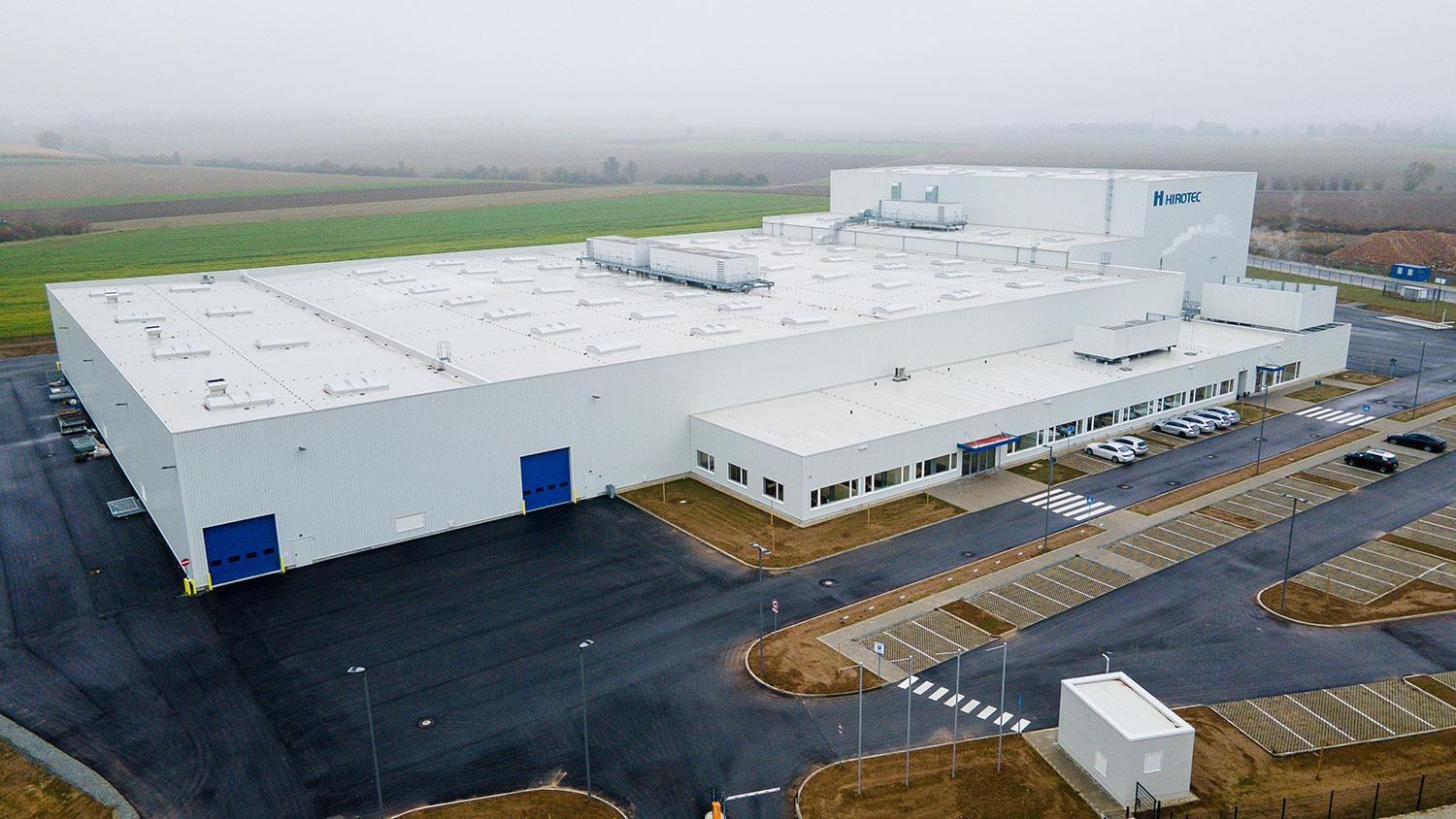 Duży, biały budynek przemysłowy z niebieskimi bramami i parkingiem, widok z góry w pochmurny dzień.