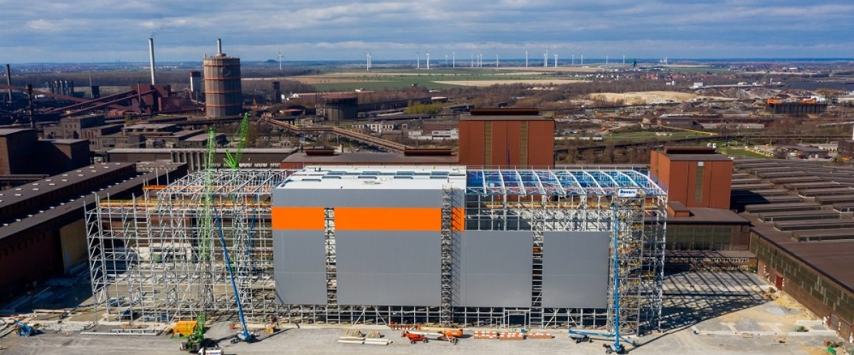 Szeroki kadr na budowę hali stalowej z częściowo zamontowanymi szarymi płytami warstwowymi z pomarańczowym pasem, widoczne rusztowania i żurawie na tle przemysłowego krajobrazu z kominami...