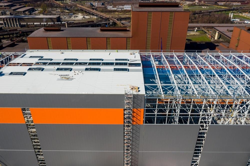 Wysoka hala w trakcie budowy z widoczną stalową konstrukcją dachu i częściowo zamontowanymi szarymi ścianami z poziomym pomarańczowym pasem. Na dachu widoczne świetliki i rusztowania, w tle...