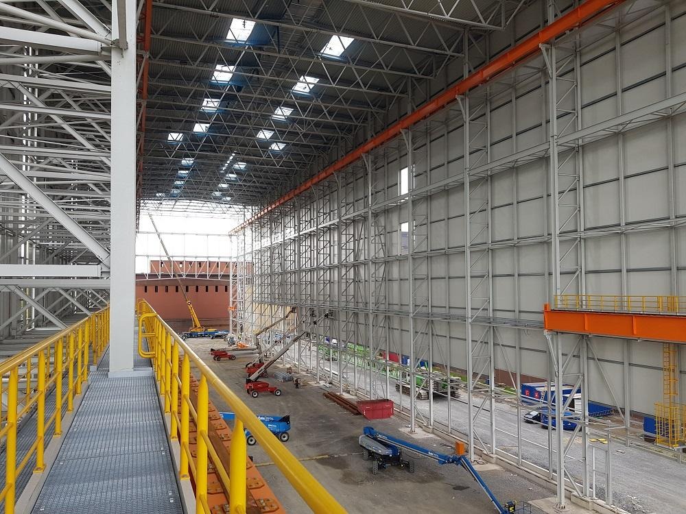 Wnętrze budowanej hali z widoczną konstrukcją stalową, jasnymi płytami ściennymi, świetlikami w dachu i sprzętem budowlanym na podłodze. Żółte barierki wzdłuż kładki dla pieszych w lewym dolnym rogu.
