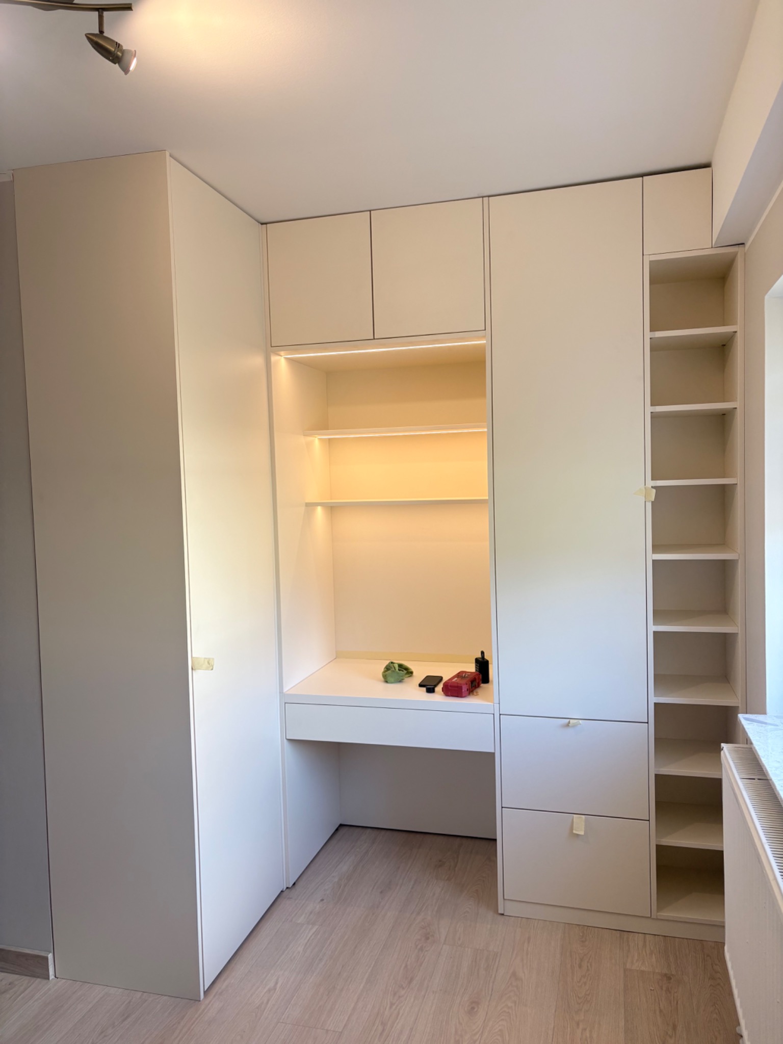 Biała szafa wnękowa z biurkiem i oświetleniem LED, widok z przodu. Na blacie biurka leżą drobne przedmioty. Minimalistyczny design, jasne wnętrze.