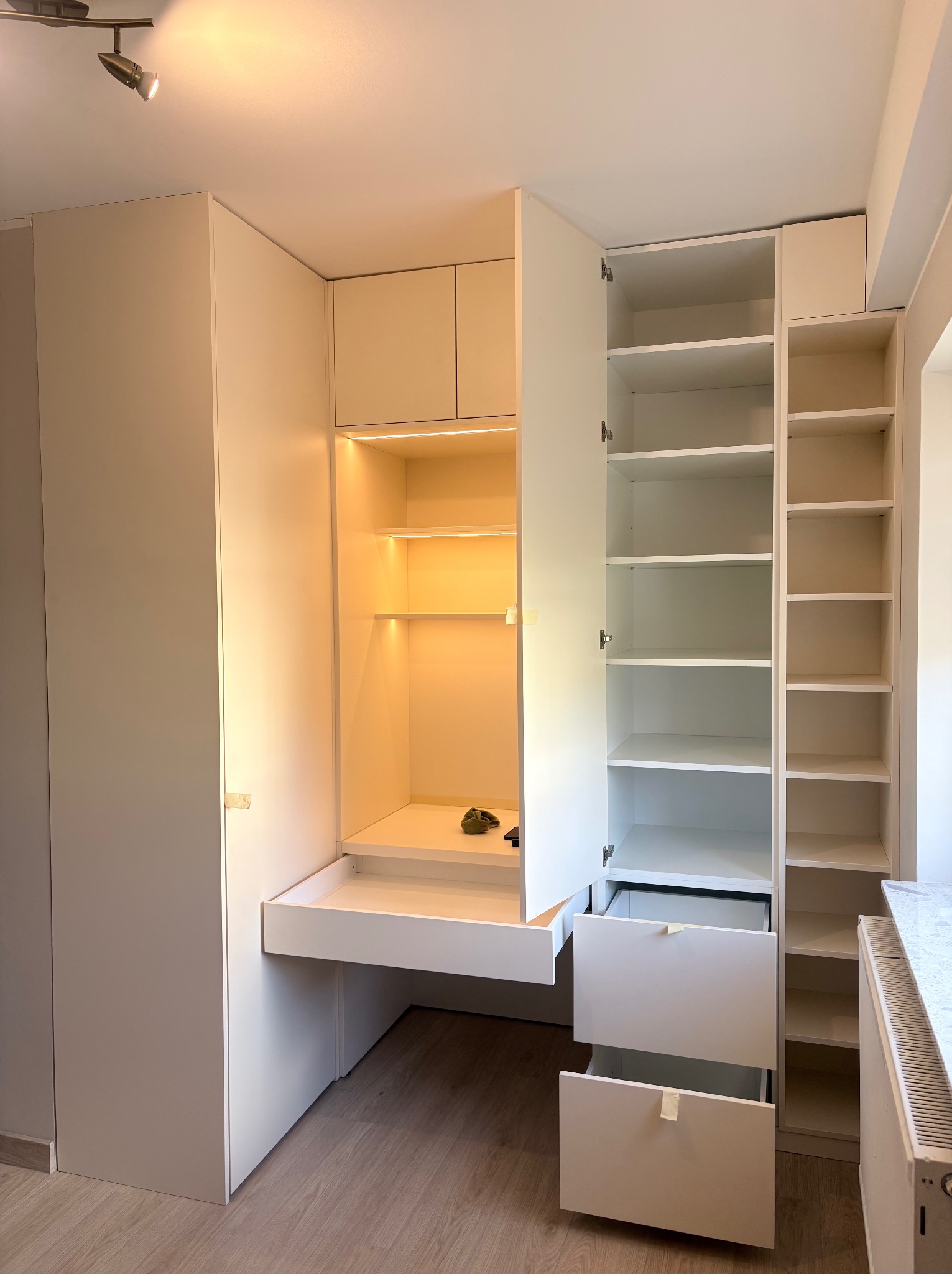 Biała szafa wnękowa z biurkiem i oświetleniem LED, otwarte drzwi i szuflady, widoczne półki. Minimalistyczny design, jasne wnętrze, drewniana podłoga.