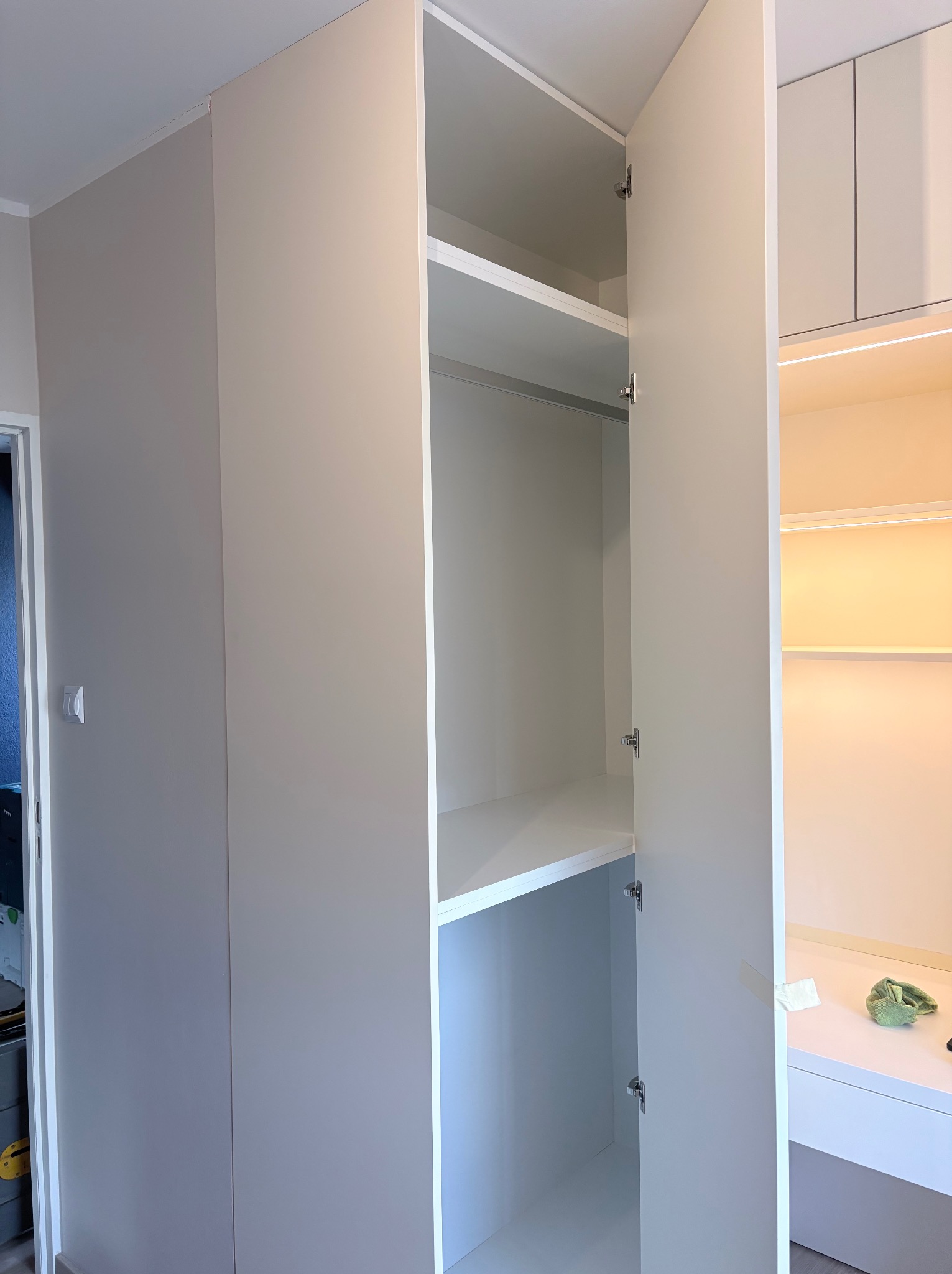 Jasna szafa wnękowa z otwartymi drzwiami, ukazująca półki i oświetlenie wewnętrzne. Minimalistyczny design, gładkie fronty, widoczne zawiasy.