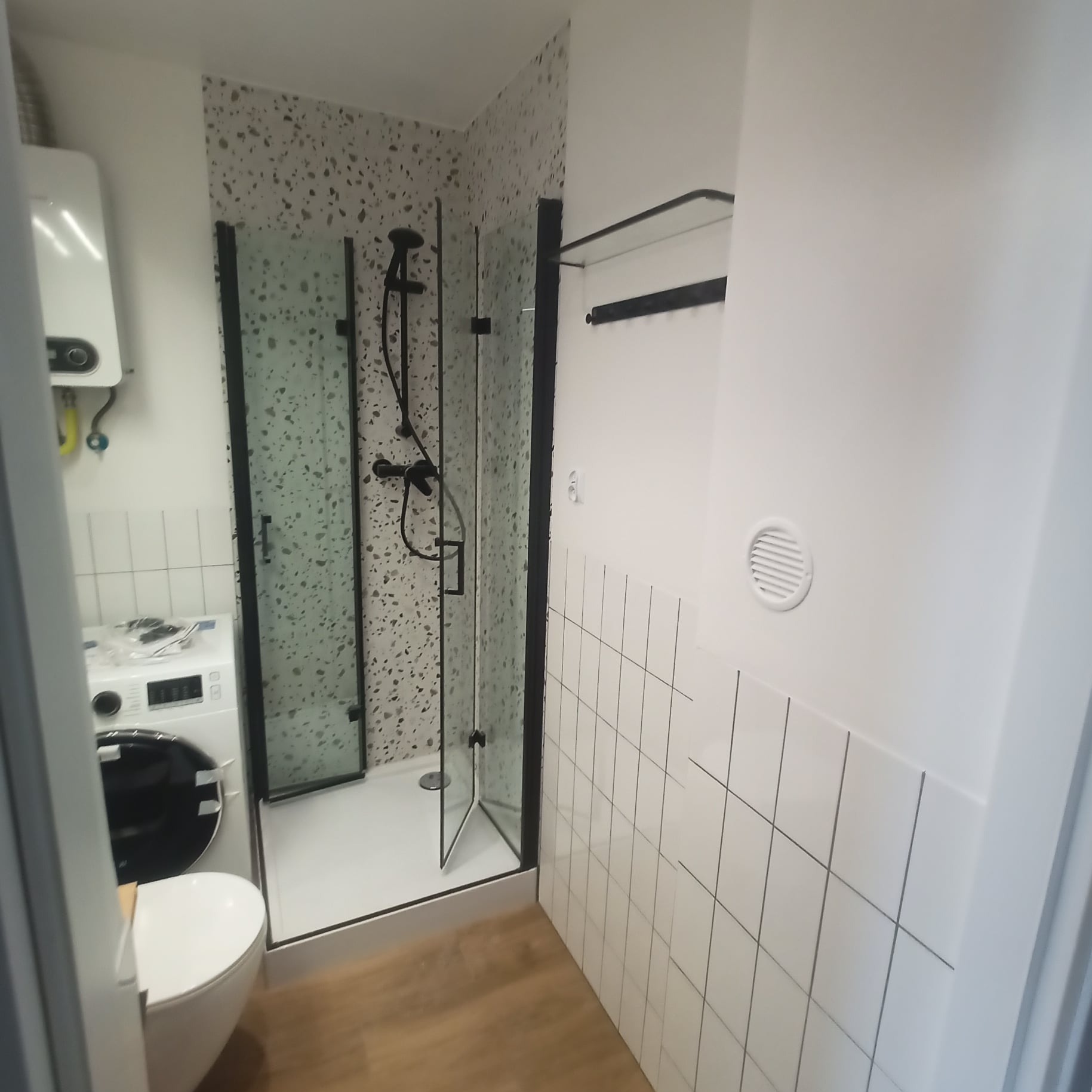 Nowoczesna łazienka z kabiną prysznicową w czarnej ramie, umywalką, pralką i bojlerem, wykończona płytkami w drobną mozaikę i białymi kaflami z czarną fugą.