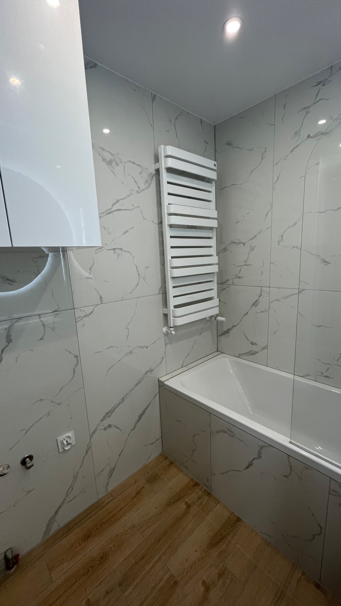 Nowoczesna łazienka z wanną obudowaną płytkami imitującymi marmur, drewnopodobną podłogą, grzejnikiem drabinkowym i białymi szafkami z połyskiem.