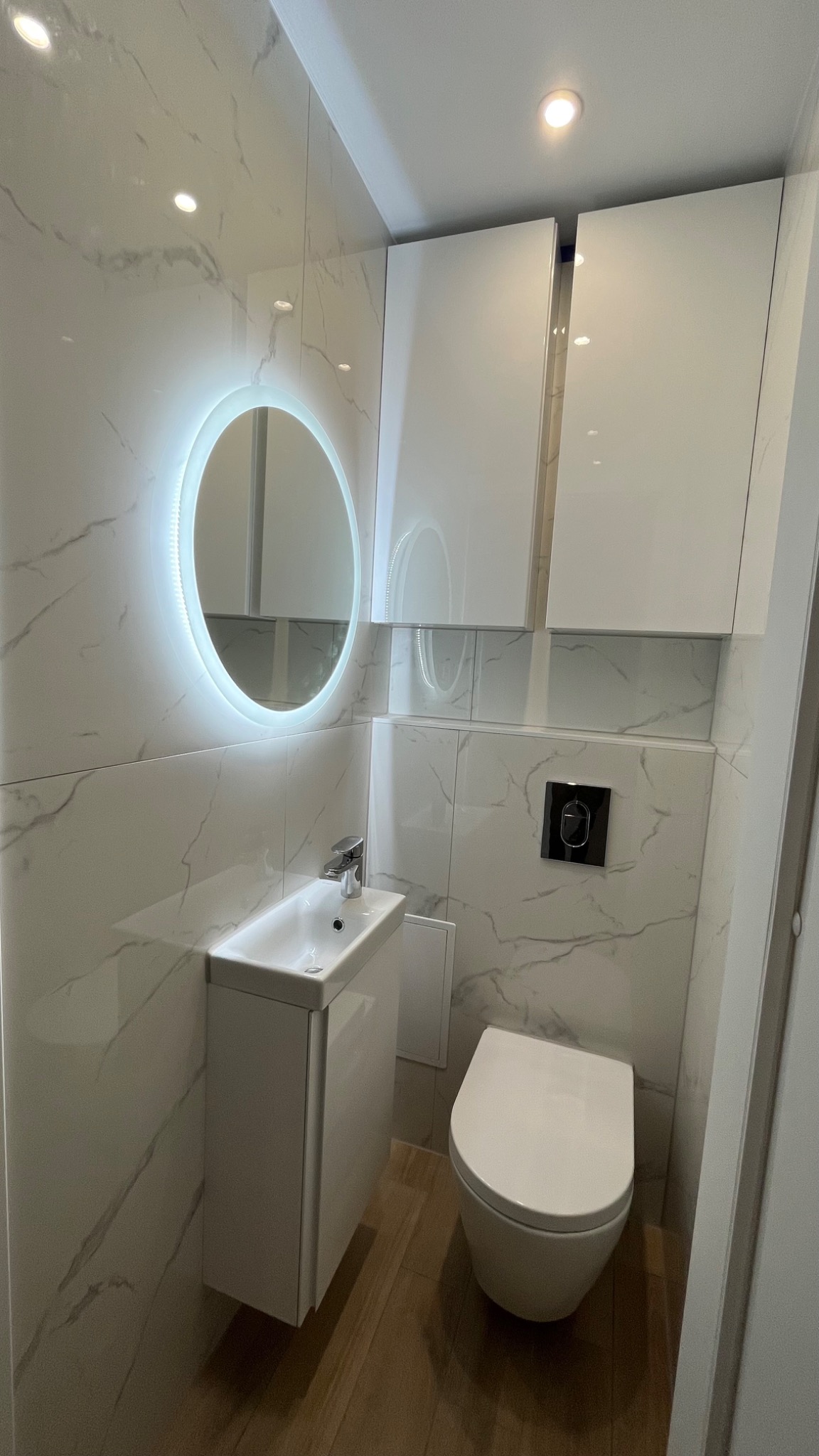 Nowoczesna łazienka z marmurowymi płytkami, podświetlanym okrągłym lustrem, wiszącą szafką umywalkową i toaletą, na podłodze panele imitujące drewno.