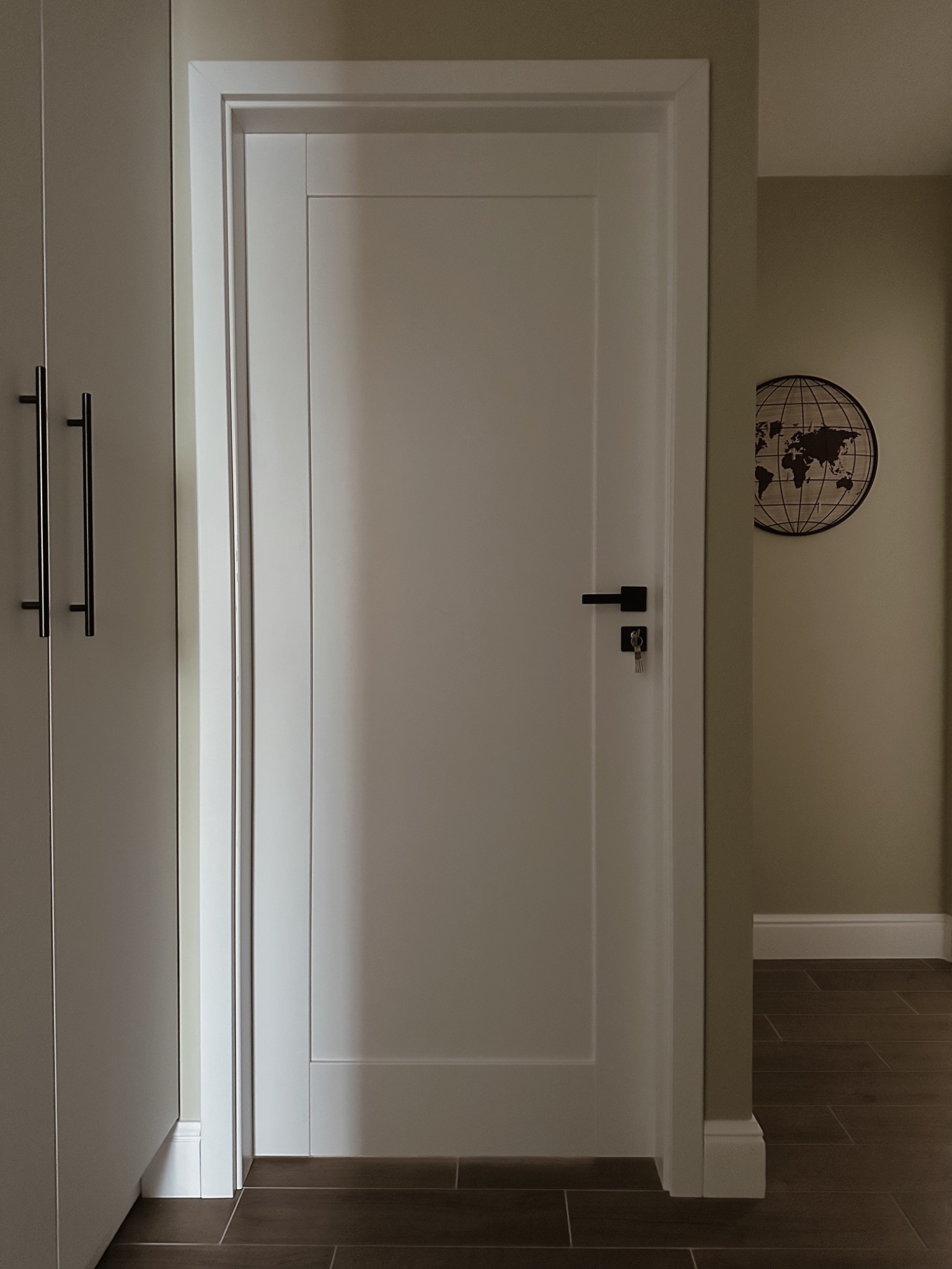 Białe, minimalistyczne drzwi wewnętrzne z czarną klamką i kluczem w zamku, widoczne na tle beżowej ściany z dekoracją w kształcie mapy świata, obok szafy wnękowej z długimi, czarnymi uchwytami.