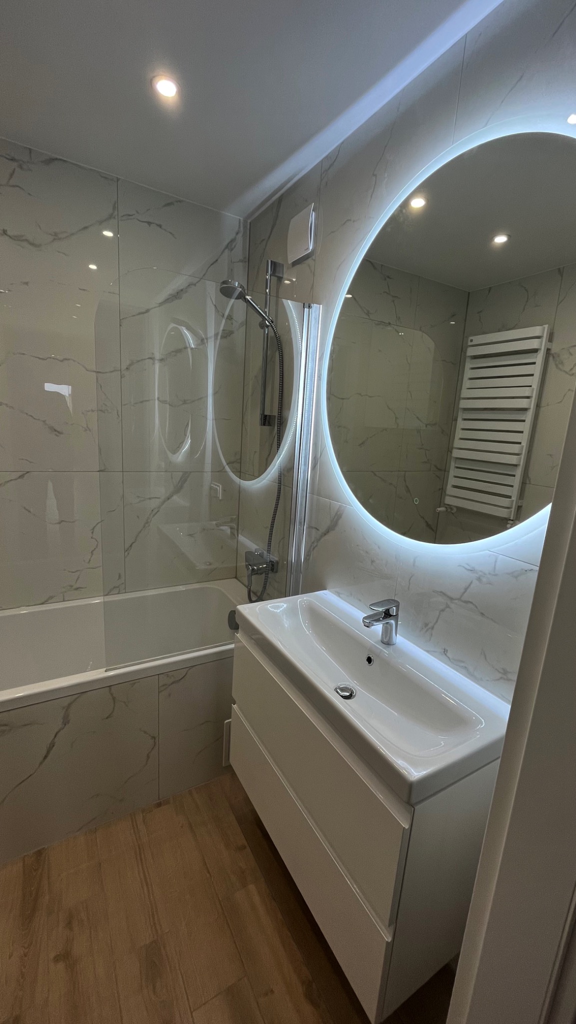 Nowoczesna łazienka z marmurowymi płytkami, podświetlanym okrągłym lustrem, białą szafką pod umywalką i drewnopodobną podłogą.