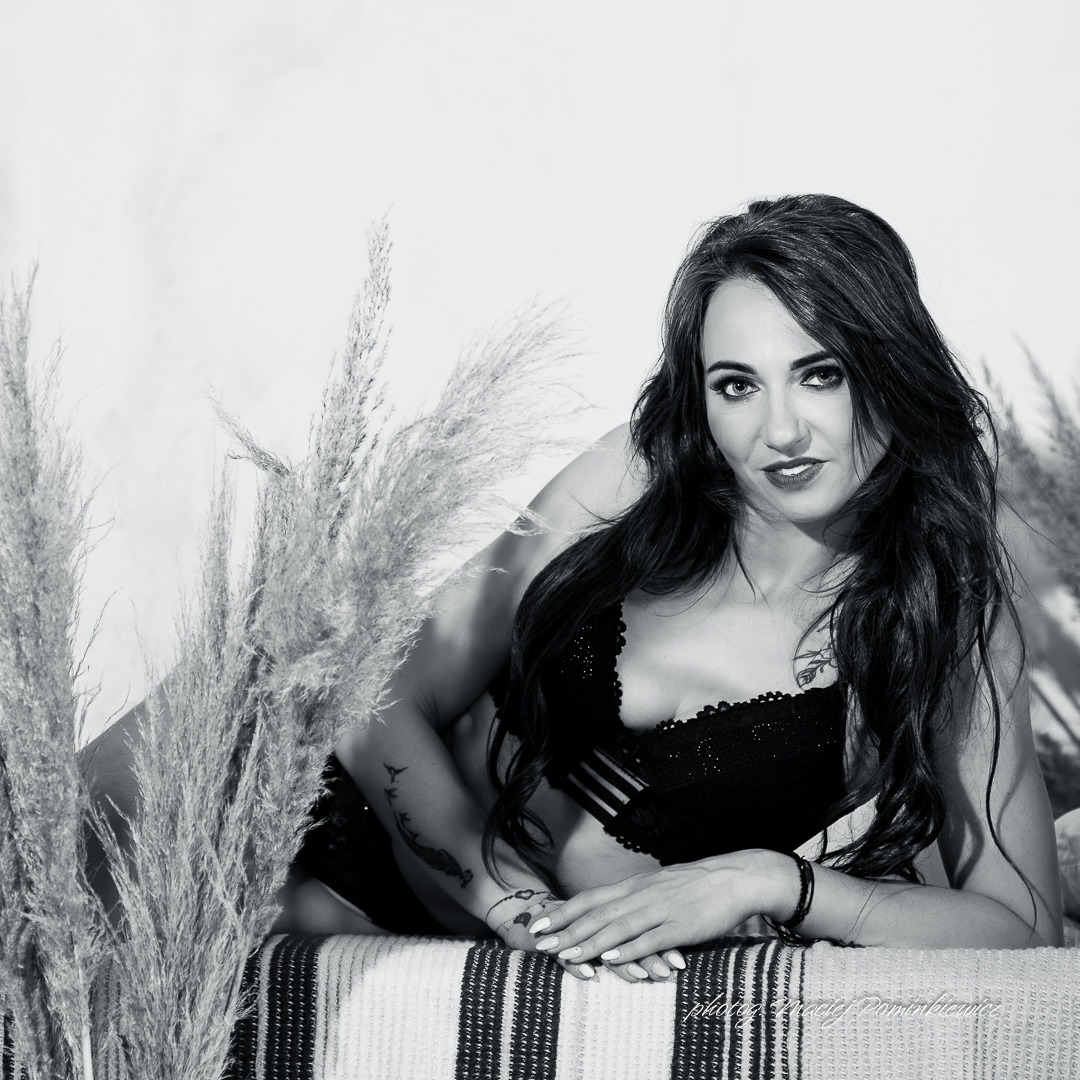 Czarno-białe ujęcie kobiety w bieliźnie leżącej na paskowanym materiale, otoczonej trawą pampasową, z tatuażami na ramieniu i nadgarstku.