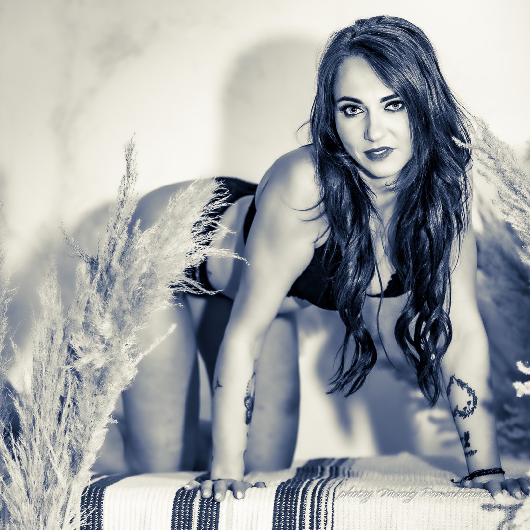 Kobieta w bieliźnie pozuje na czarno-białym zdjęciu, opierając się na rękach na wzorzystej tkaninie, z puszystymi trawami po bokach i tatuażami na ramionach.