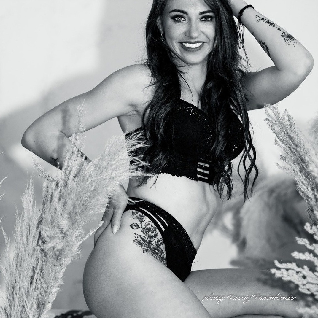 Czarno-białe zdjęcie uśmiechniętej kobiety w bieliźnie, z tatuażami na ramieniu i udzie, pozującej na tle pampasowej trawy.
