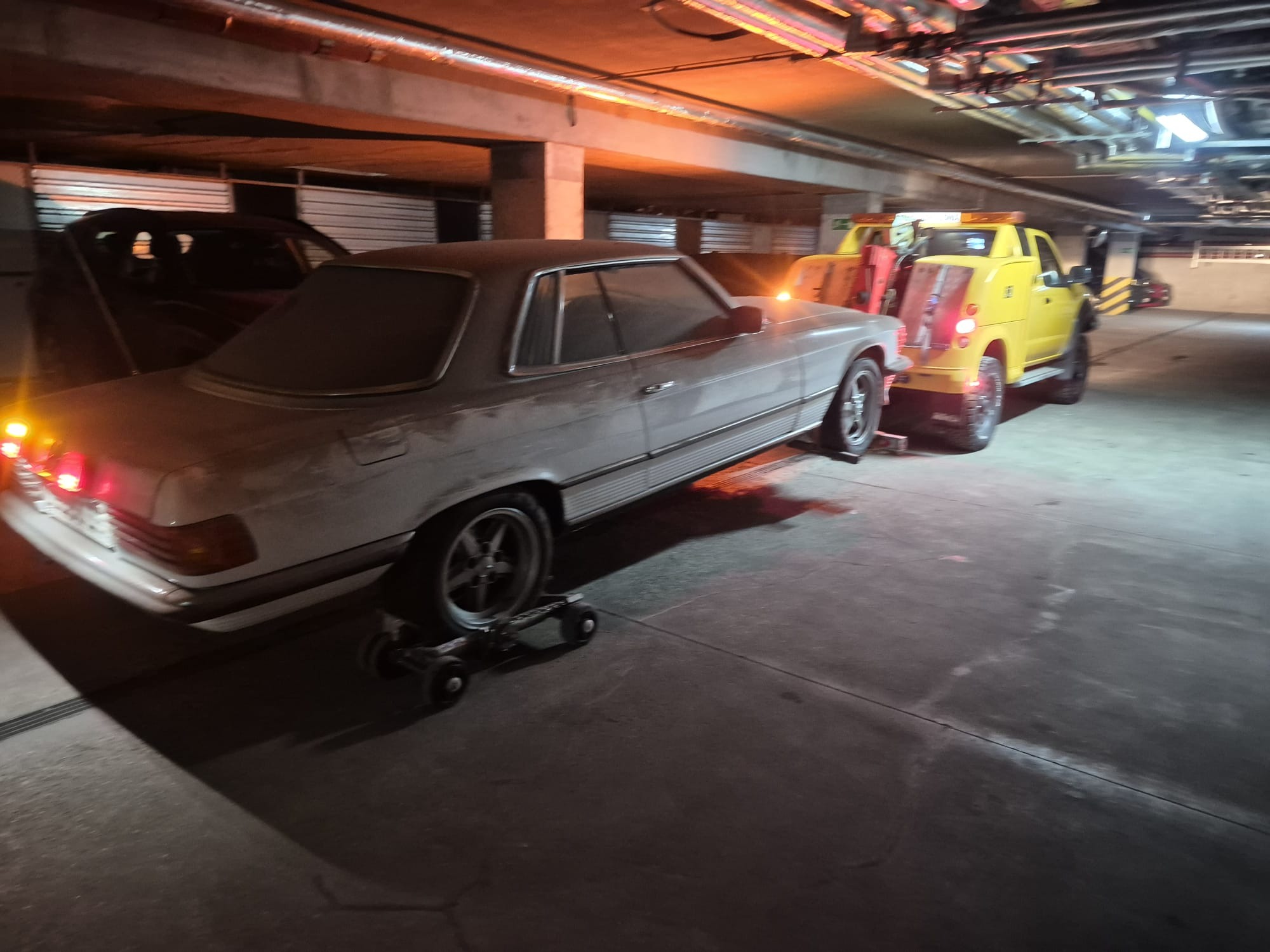Żółta pomoc drogowa holuje zakurzone, srebrne auto na wózkach w garażu podziemnym. W tle widoczne inne pojazdy i instalacje pod sufitem.