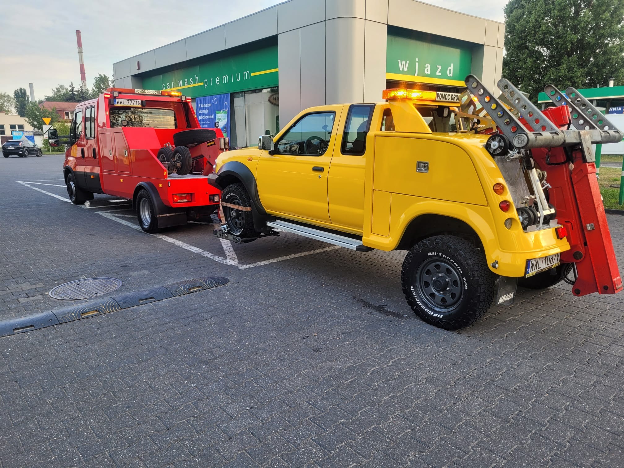 Żółta laweta holuje uszkodzony pojazd na parkingu stacji benzynowej. Widoczna czerwona laweta z napisem 'Pomoc Drogowa'.