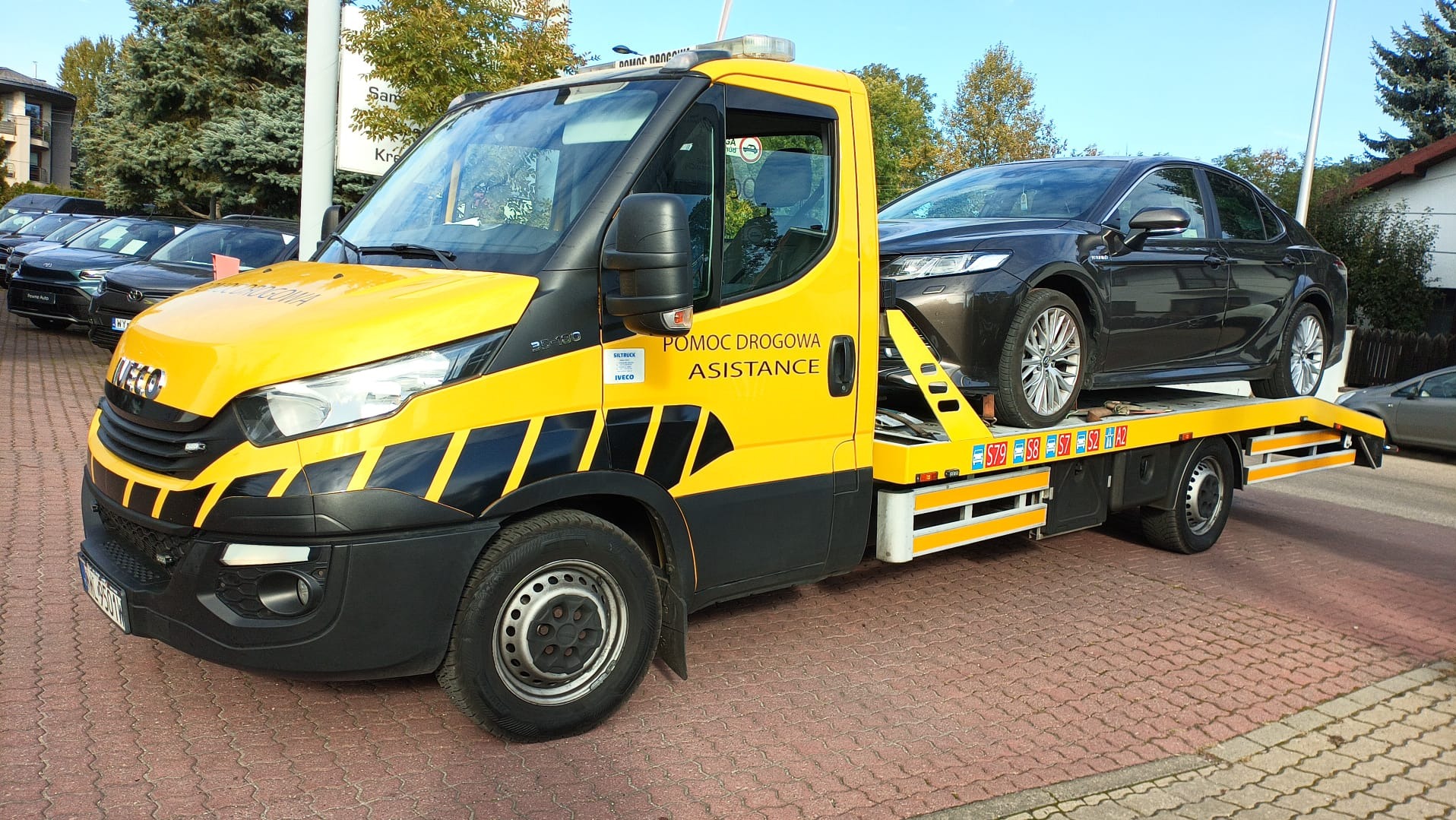 Żółta autolaweta Iveco z czarnymi pasami transportuje ciemny samochód osobowy. Widoczny napis 'Pomoc Drogowa Assistance'.