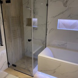 Robert Fritz - Nowoczesna łazienka z kabiną prysznicową walk-in z czarnymi okuciami, białą wanną z podświetleniem LED, marmurowymi płytkami ściennymi i drewnopodobną podłogą.