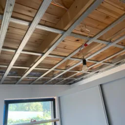 Stelaż metalowy podwieszanego sufitu z drewnianym poszyciem, widoczne przewody elektryczne i żar&oacute;wka, fragment okna i ściany w pomieszczeniu.