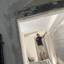 Robert Fritz - Mężczyzna na aluminiowej drabinie szpachluje sufit w trakcie remontu, widoczne niedoskonałości i materiały budowlane, zabezpieczona folią ściana oraz fragment zbrojenia sufitu.