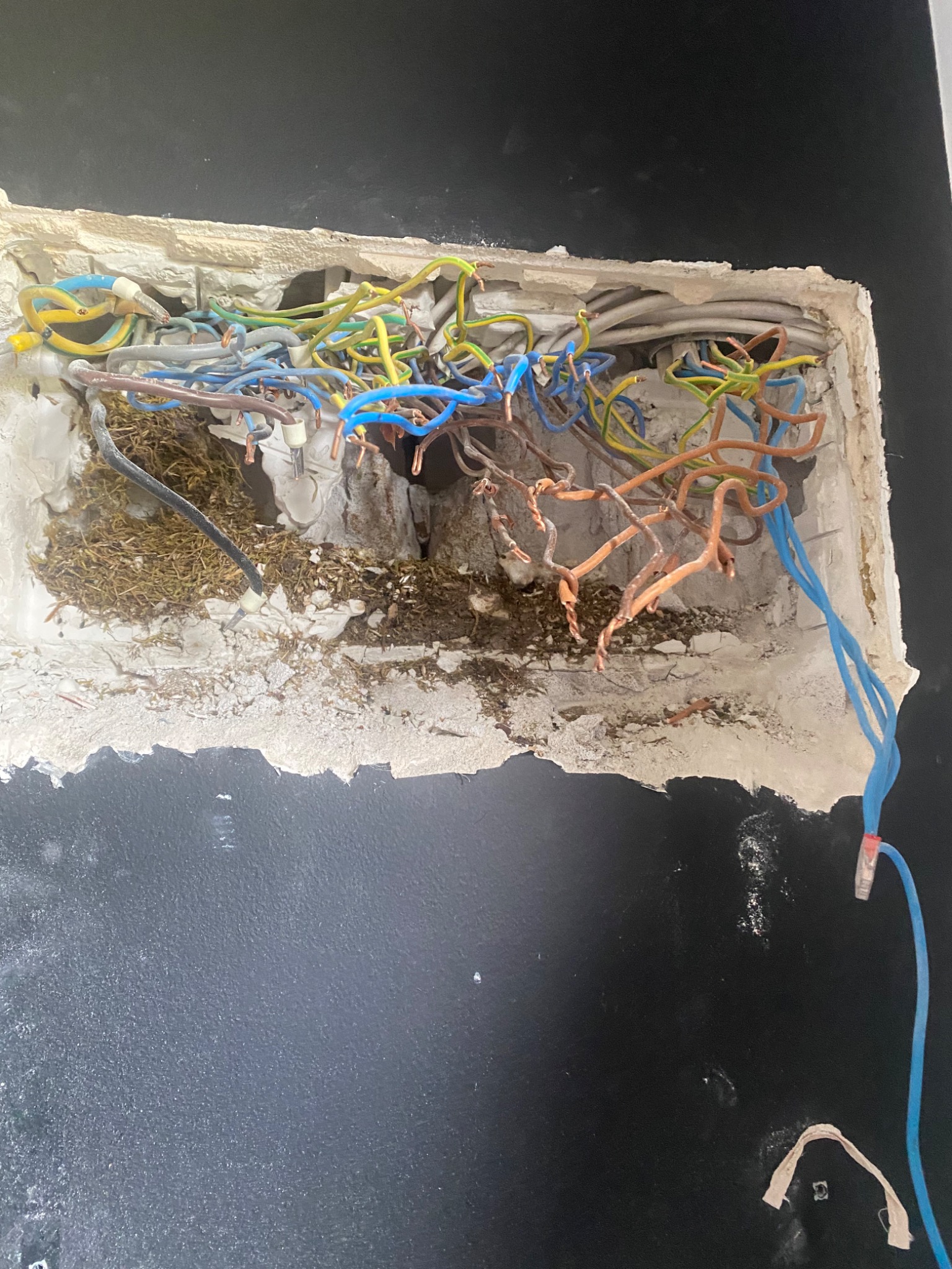 Otwór w ścianie z widocznymi przewodami elektrycznymi o różnych kolorach (niebieski, żółty, zielony, brązowy), częściowo zabezpieczonymi izolacją, z widocznymi fragmentami gruzu i resztek...