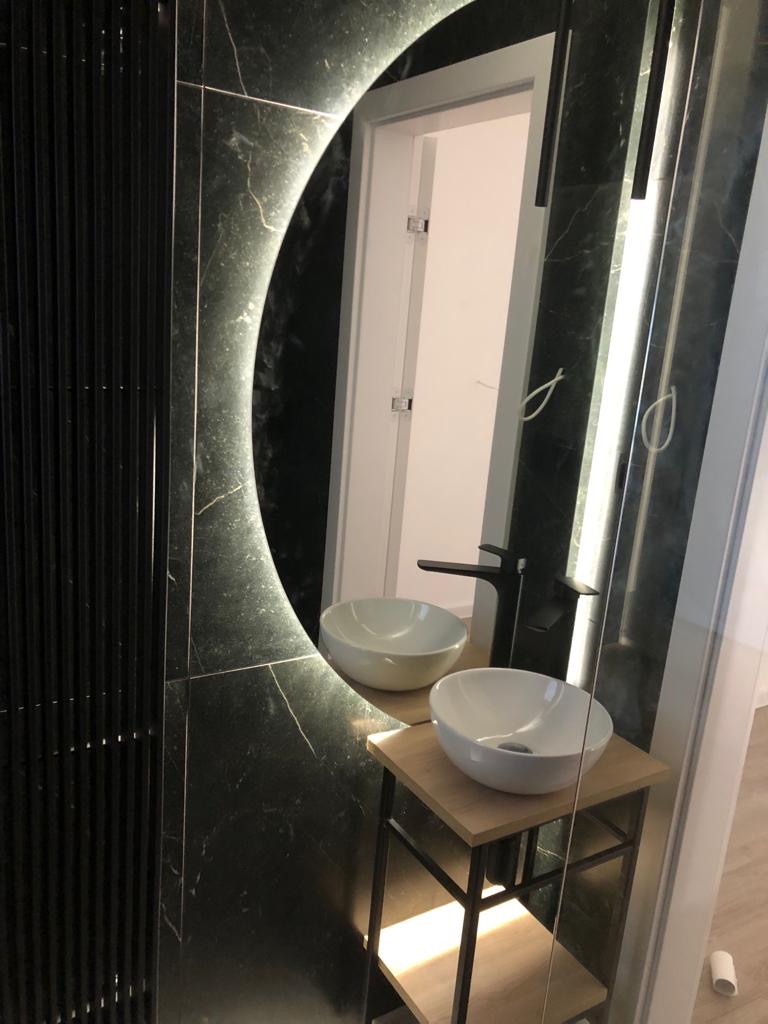 Nowoczesna łazienka z czarnymi marmurowymi płytkami, okrągłym lustrem z podświetleniem LED i dwiema białymi umywalkami nablatowymi na drewnianym regale z czarną ramą.