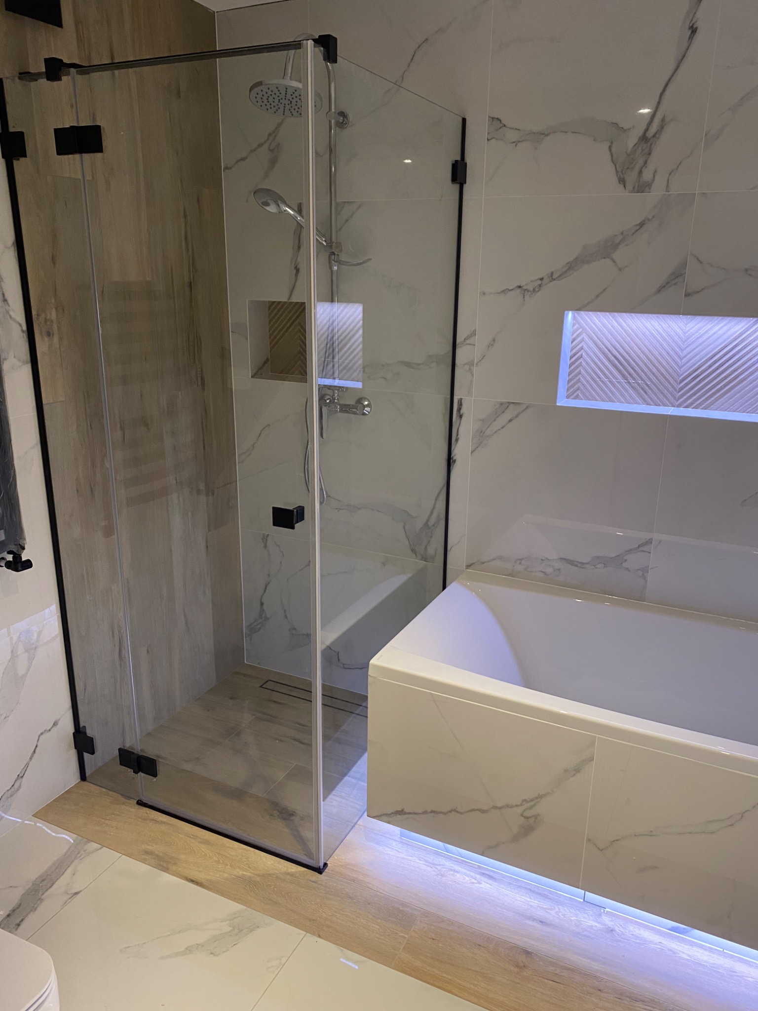Nowoczesna łazienka z kabiną prysznicową walk-in z czarnymi okuciami, białą wanną z podświetleniem LED, marmurowymi płytkami ściennymi i drewnopodobną podłogą.