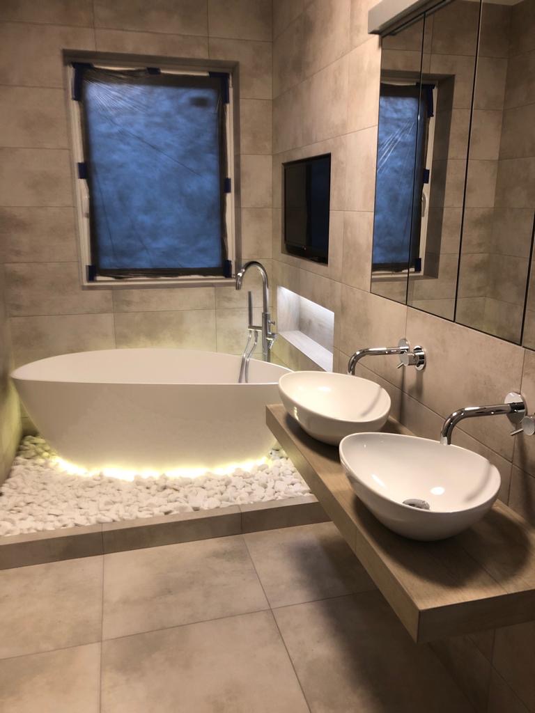 Nowoczesna łazienka z owalną wanną podświetloną białymi kamieniami, dwie umywalki nablatowe na drewnianym blacie, lustro w szafce i telewizor w ścianie wyłożonej płytkami w odcieniach beżu.