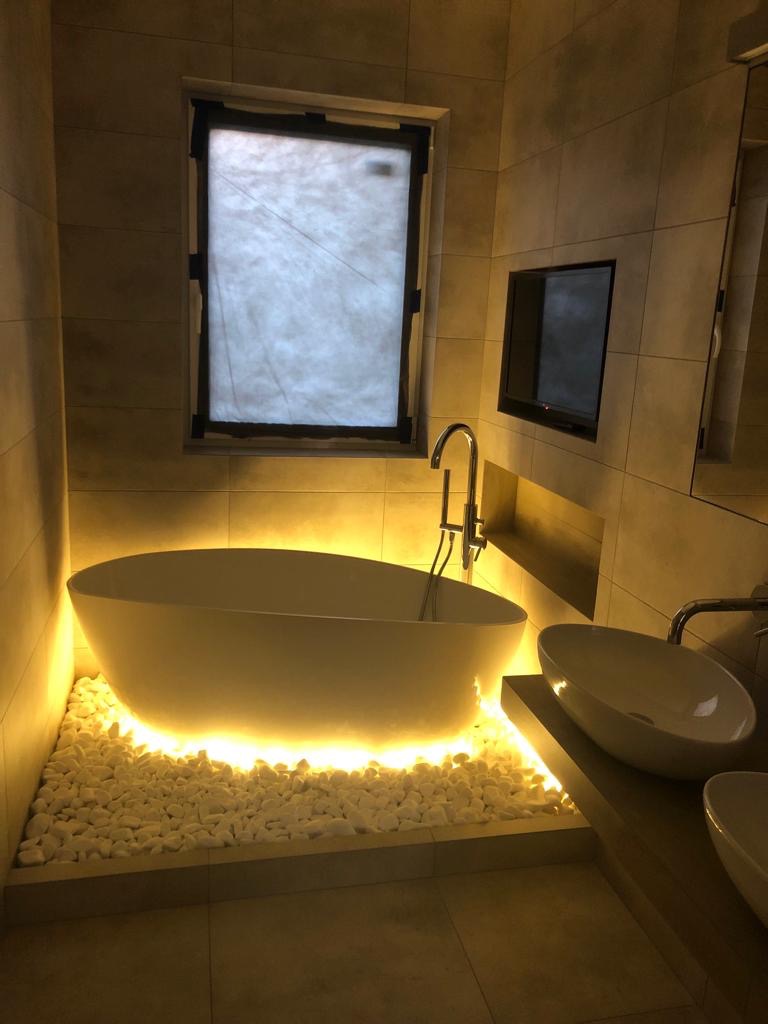Nowoczesna łazienka z wolnostojącą wanną podświetloną od dołu białymi kamieniami, minimalistyczna armatura i telewizor wbudowany w ścianę.