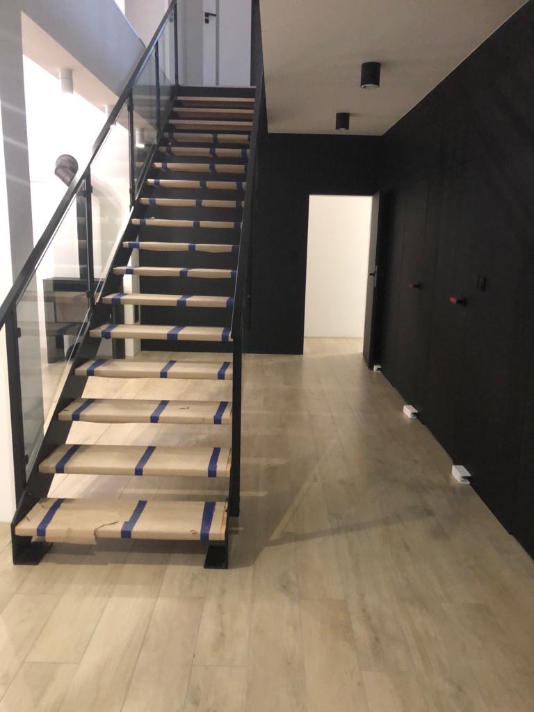 Nowoczesne schody drewniane z czarną balustradą, stopnie zabezpieczone papierem i taśmą malarską, jasna podłoga, czarna ściana w tle.