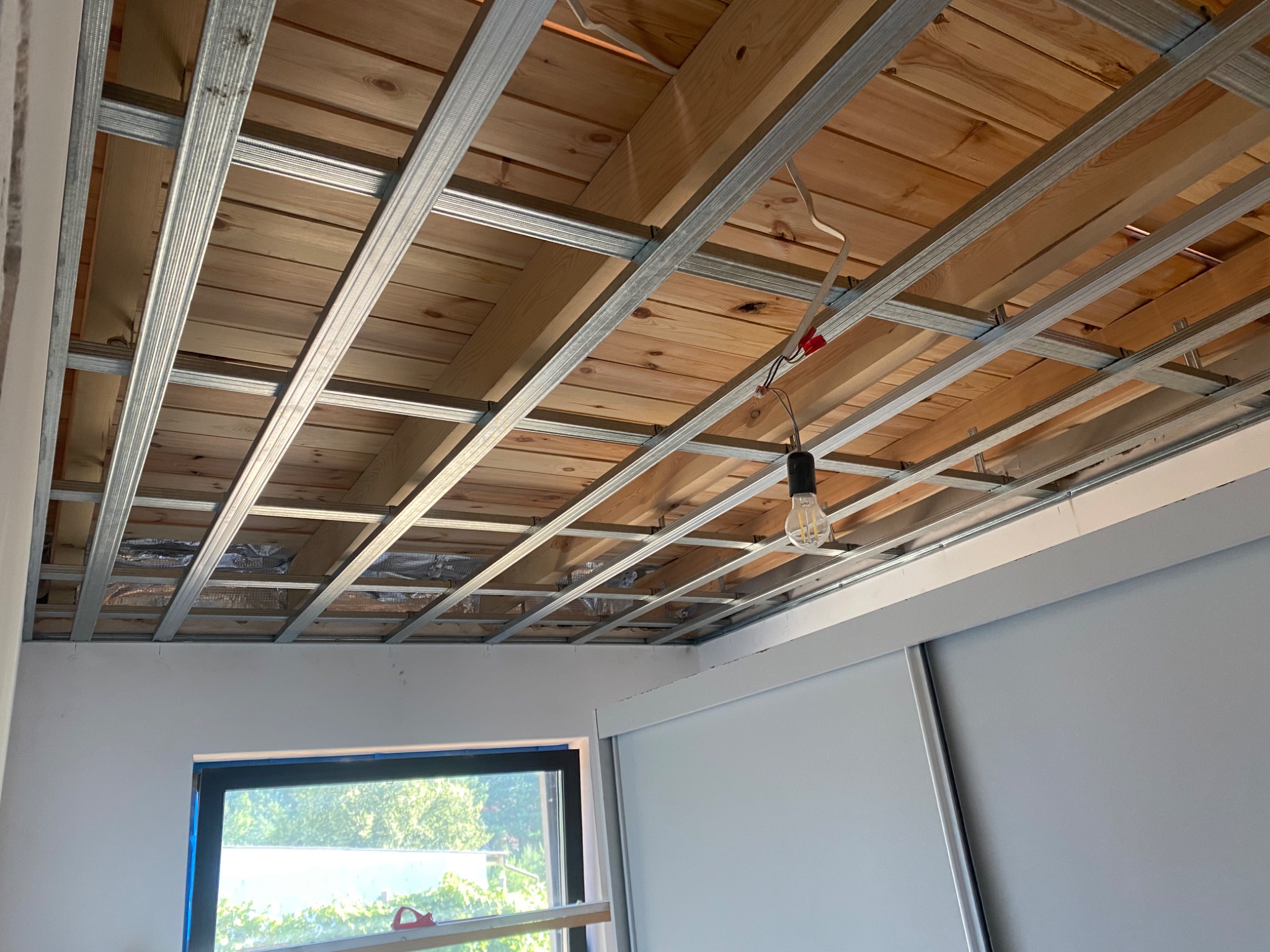 Stelaż metalowy podwieszanego sufitu z drewnianym poszyciem, widoczne przewody elektryczne i żarówka, fragment okna i ściany w pomieszczeniu.