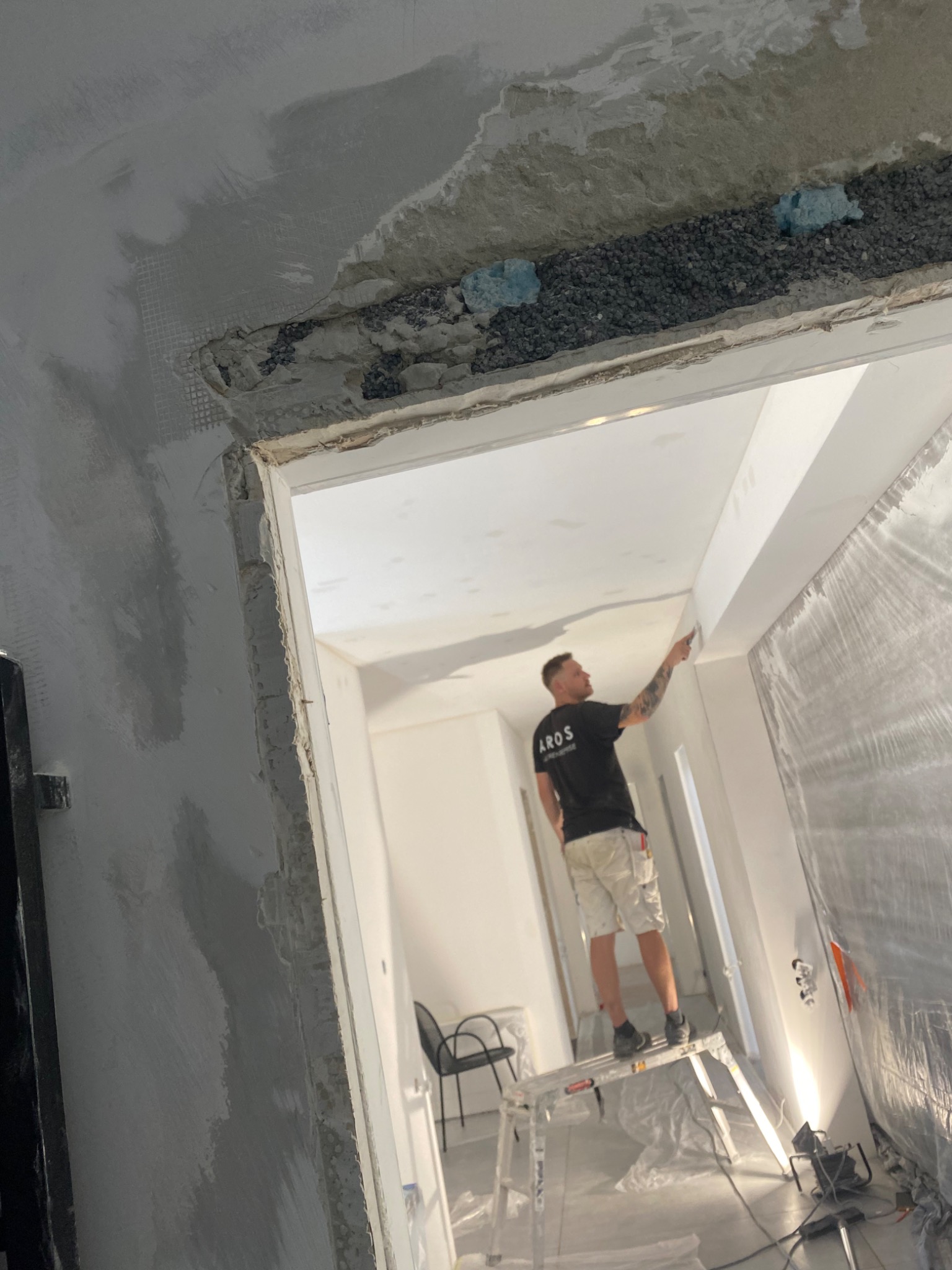 Mężczyzna na aluminiowej drabinie szpachluje sufit w trakcie remontu, widoczne niedoskonałości i materiały budowlane, zabezpieczona folią ściana oraz fragment zbrojenia sufitu.