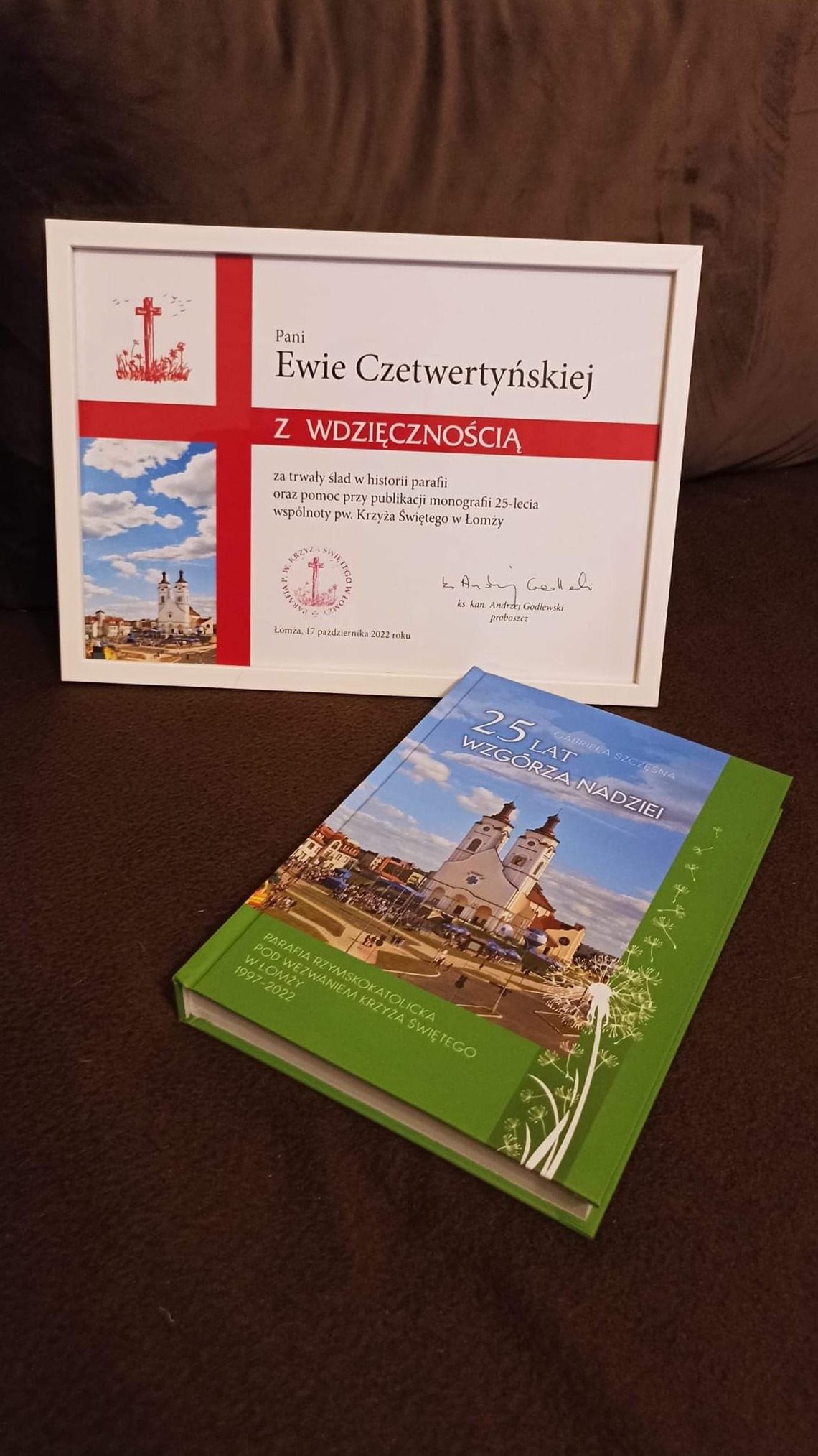 Oprawiona w ramkę dedykacja dla Ewy Czetwertyńskiej z podziękowaniem za wkład w historię parafii i pomoc w publikacji monografii, obok leży książka o 25-leciu parafii rzymskokatolickiej...