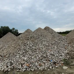Kupa kamieni i gruzu z fragmentami cegieł ułożona w stożki na placu budowy, z widocznymi drzewami i pochmurnym niebem w tle.