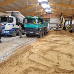 Wnętrze hali z dwoma samochodami ciężarowymi i koparką podczas utwardzania podłoża piaskiem i gruzem, widoczni dwaj pracownicy w odzieży roboczej.