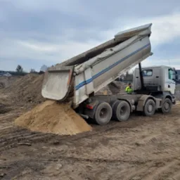 Ciężarówka z podniesioną skrzynią wyładowuje jasny materiał sypki na placu budowy, w tle hałdy ziemi i zabudowania.